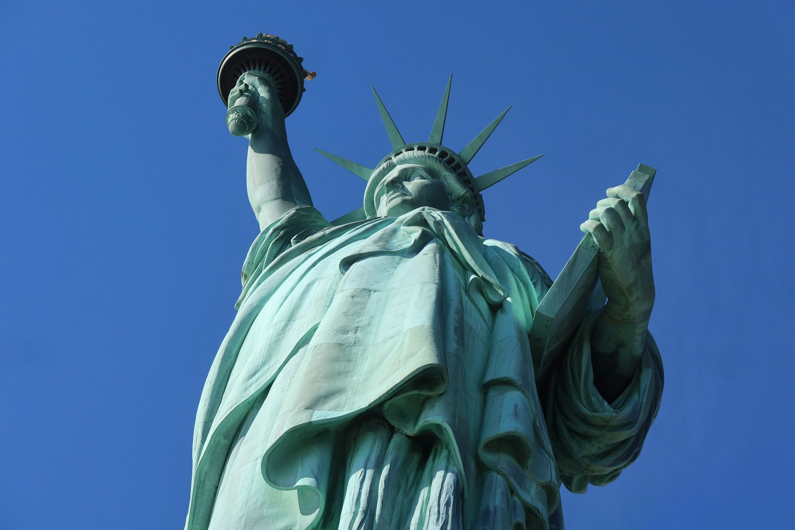 Low angle photo looking up at the Statue of Liberty against a clear blue sky. The photo emphasizes her tablet, crown, and torch.